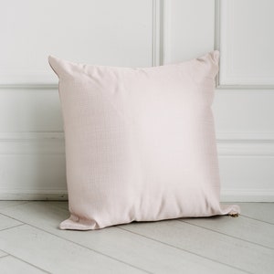 May include: A pale pink pillow with a subtle textured surface. The pillow is lying on a white wood floor.