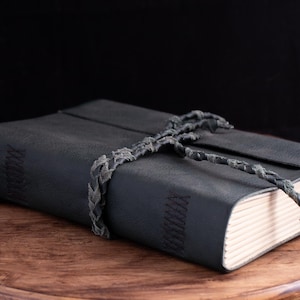 Könnte beinhalten: Ein dunkelgraues Lederjournal mit geflochtenem Kordelverschluss. Das Journal ist geschlossen und liegt auf einer Holzoberfläche. Die Seiten sind sichtbar und der Einband hat Ziernähte.