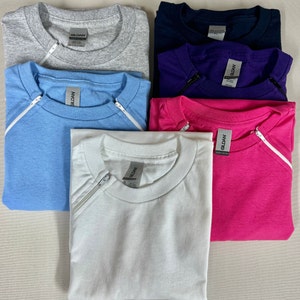 May include: Six t-shirts in different colours, including grey, blue, purple, pink, and white. Each shirt has a zipper on the shoulder and a Gildan label.