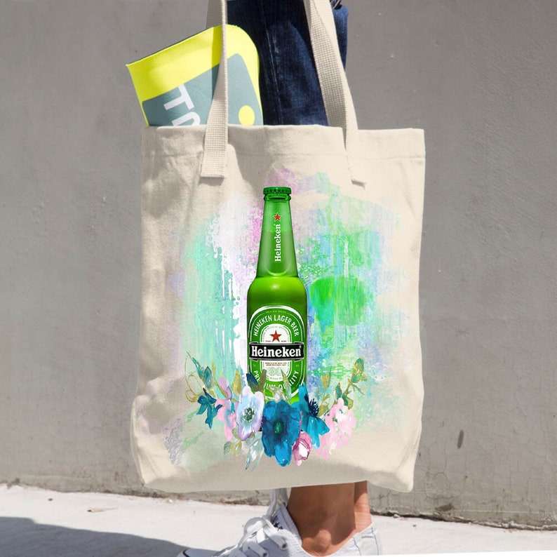 May include: A beige tote bag featuring a green Heineken beer bottle with a floral design. The bottle is centered on a colorful abstract background. The bag is carried by a person wearing blue jeans and white sneakers.