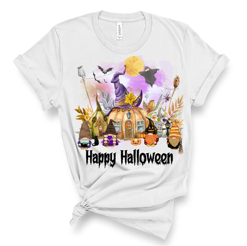 Pu&ograve; includere: Una maglietta bianca con un'illustrazione colorata di un villaggio di gnomi con una casa a forma di zucca, un cappello da strega e un fantasma. Il testo "Happy Halloween" &egrave; stampato sotto l'illustrazione.