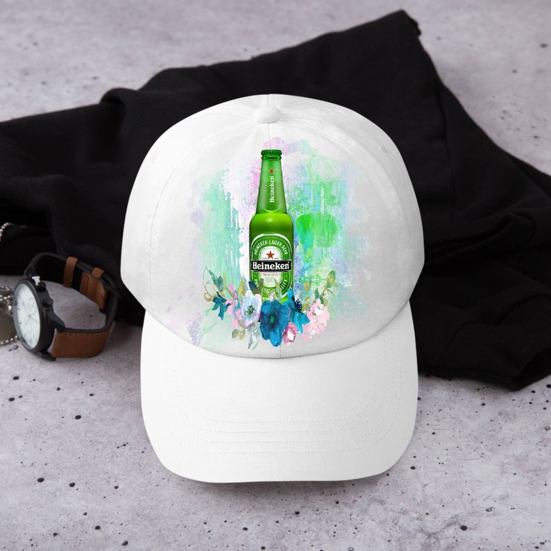 May include: White baseball cap featuring a green Heineken beer bottle design with floral accents. The cap has a curved brim and a classic six-panel construction. The design includes a watercolor-style background in shades of green, blue, and pink.