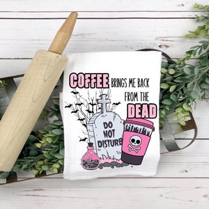 以下が含まれることがあります： 白いキッチンクロスに、ピンクと黒のグラフィックが描かれています。コーヒーカップにはドクロとクロスボーン、墓石には「邪魔しないで」の文字、そして墓石の周りをコウモリが飛んでいます。テキストには「コーヒーは私を死者の世界から連れ戻してくれる」と書いてあります。