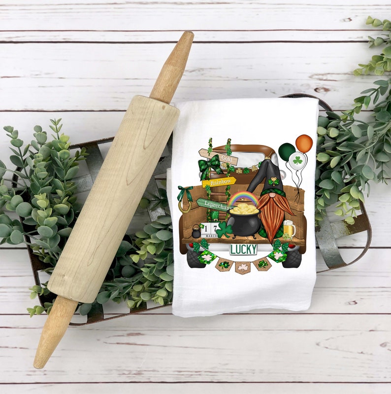 May include: A white kitchen towel with a green, brown, and gold illustration of a truck with a gnome sitting in the back. The truck is decorated with shamrocks, gold coins, and a pot of gold. The truck has a license plate that says "LUCKY".