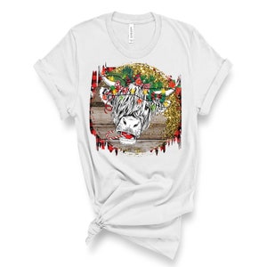 May include: White t-shirt featuring a Christmas-themed design of a Highland cow with a red candy cane and a wreath of lights and bows on its head. The design is set against a woodgrain background with gold glitter.