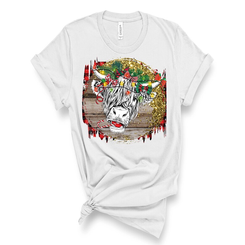 May include: White t-shirt featuring a Christmas-themed design of a Highland cow with a red candy cane and a wreath of lights and bows on its head. The design is set against a woodgrain background with gold glitter.