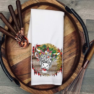May include: White kitchen towel featuring a festive Highland cow design. The cow is wearing a wreath of holly and has a red nose. The background is a rustic woodgrain with gold glitter.