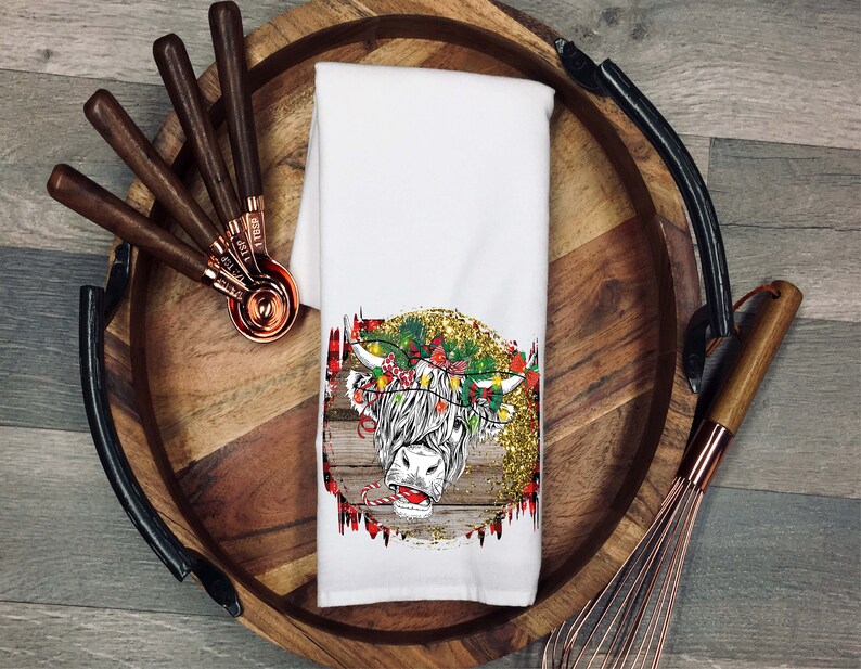 May include: White kitchen towel featuring a festive Highland cow design. The cow is wearing a wreath of holly and has a red nose. The background is a rustic woodgrain with gold glitter.