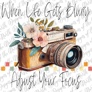 May include: A vintage camera with a lens and a floral arrangement of pink and white flowers. The text "When life gets blurry adjust your focus" is written in black cursive.