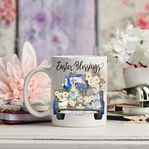 Peut inclure: Une tasse &agrave; caf&eacute; en c&eacute;ramique blanche avec un motif de camionnette bleue. Le camion est rempli de d&eacute;corations de P&acirc;ques, dont un nichoir, des fleurs et une croix. Le texte "Easter Blessings" est &eacute;crit sur la tasse.