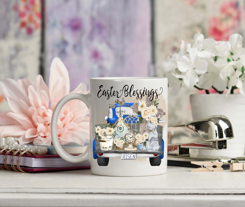 Peut inclure: Une tasse &agrave; caf&eacute; en c&eacute;ramique blanche avec un motif de camionnette bleue. Le camion est rempli de d&eacute;corations de P&acirc;ques, dont un nichoir, des fleurs et une croix. Le texte "Easter Blessings" est &eacute;crit sur la tasse.