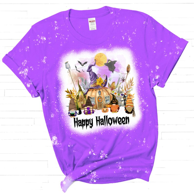 Pu&ograve; includere: Maglietta viola con un designo schizzato di candeggina che presenta un'illustrazione ad acquerello di gnomi, una casa a forma di zucca e un cappello da strega. Il testo "Happy Halloween" &egrave; stampato sotto l'illustrazione.