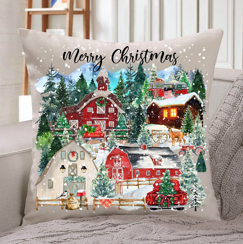 May include: A beige throw pillow with a watercolor illustration of a snowy winter village scene. The pillow features red barns, green trees, and a red truck with a Christmas tree in the back. The text "Merry Christmas" is printed in black at the top of the pillow.