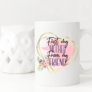 Peut inclure: Une tasse en c&eacute;ramique blanche avec un motif de c&oelig;ur dor&eacute; et le texte "First my Mother forever my Friend". Le c&oelig;ur est rempli d'un motif aquarelle rose et d'une illustration florale.