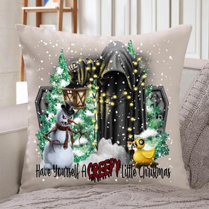 Op de afbeelding: Een wit kussen met een feestelijke kerstsc&egrave;ne met een zwarte gekapte figuur, een sneeuwpop met een dreigende grijns, een rubberen eend en een grafsteen. De tekst "Have Yourself A Creepy Little Christmas" is op het kussen gedrukt.