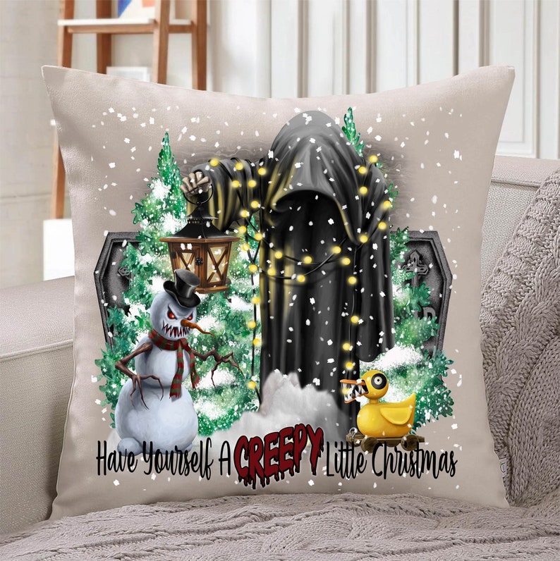Op de afbeelding: Een wit kussen met een feestelijke kerstsc&egrave;ne met een zwarte gekapte figuur, een sneeuwpop met een dreigende grijns, een rubberen eend en een grafsteen. De tekst "Have Yourself A Creepy Little Christmas" is op het kussen gedrukt.