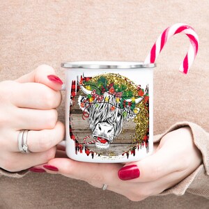 May include: White enamel mug with a Christmas-themed design featuring a Highland cow with a candy cane in its mouth. The cow is wearing a wreath of holly and lights. The background is a rustic woodgrain with gold glitter.