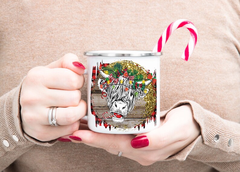 May include: White enamel mug with a Christmas-themed design featuring a Highland cow with a candy cane in its mouth. The cow is wearing a wreath of holly and lights. The background is a rustic woodgrain with gold glitter.