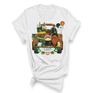 May include: A white t-shirt with a graphic of a brown truck with a gnome, a pot of gold, and a rainbow. The truck has a license plate that says "LUCKY" and the gnome is wearing a green hat. The truck is decorated with shamrocks and the words "Gold", "Rainbow", and "Leprechaun".