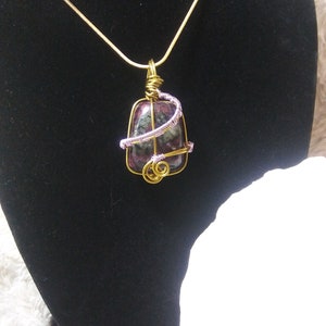 May include: A gold chain necklace with a pendant featuring a green, pink, and black stone wrapped in gold and purple wire.