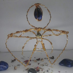 Puede incluir: Una corona de alambre dorado con una piedra preciosa azul y una piedra preciosa verde más pequeña. La corona está sobre una superficie blanca con flores y hojas secas.