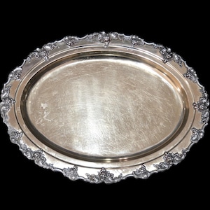 Puede incluir: Bandeja ovalada plateada con borde decorativo de uvas y hojas. La bandeja tiene una superficie ligeramente rayada y un fondo oscuro. Una pieza clásica para servir alimentos o exhibir objetos.