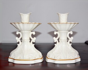 May include: Pair of white ceramic candle holders with gold trim. Each holder features a fluted top, ornate detailing, and a decorative base. The candle holders are approximately 6 inches tall.