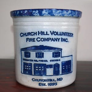 May include: A white ceramic jar with a blue glaze. The jar features a blue and white image of a fire station with the text "Church Hill Volunteer Fire Company Inc." and "Church Hill, MD Est. 1895".