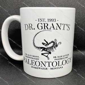 May include: White ceramic mug with a black graphic of a dinosaur skeleton. The text on the mug reads "EST. 1993 - DR. GRANT'S PALEONTOLOGY SNAKEWATER - MONTANA".