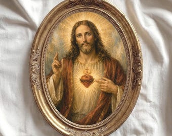 May include: An oval-shaped wall art piece featuring a portrait of a figure with long brown hair and a beard, wearing a red robe. The figure has a glowing heart on the chest and is framed in an ornate, antique-style gold-colored metal frame.