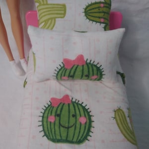 May include: A white doll bed with a pink headboard and a white and pink cactus print bedding set. The bedding set includes a fitted sheet, a flat sheet, and two pillowcases. The pillowcases have a white background with pink polka dots and a cactus with a pink bow on it. The fitted sheet and flat sheet have a white background with pink stripes and a cactus with a pink bow on it.