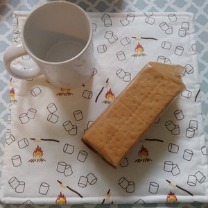 May include: A white mug with a white background placemat featuring a pattern of marshmallows and campfires. A graham cracker is on top of the placemat.