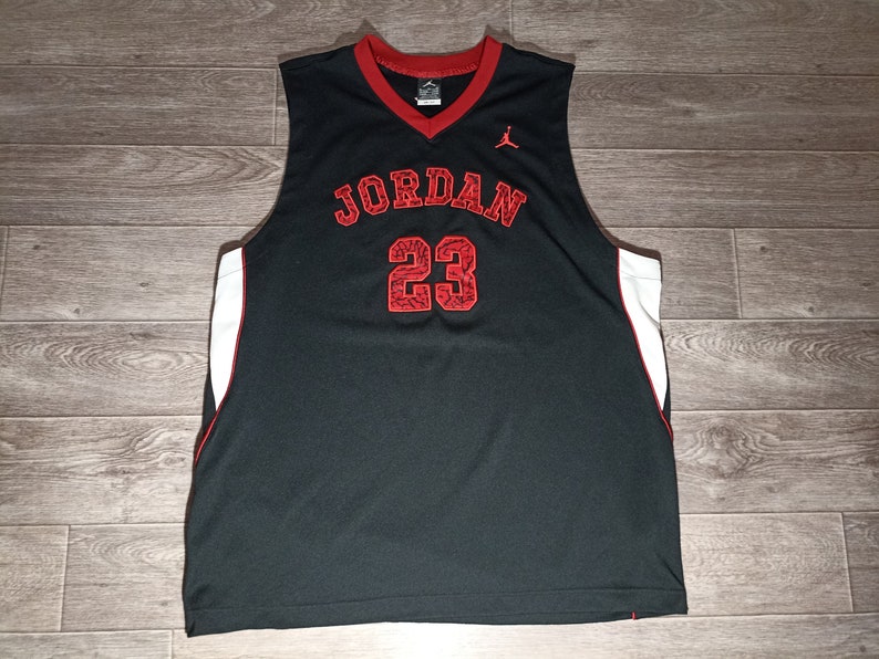 Puede incluir: Camiseta de baloncesto negra con detalles rojos y blancos. La camiseta tiene "JORDAN" y "23" impresos en la parte delantera en rojo.
