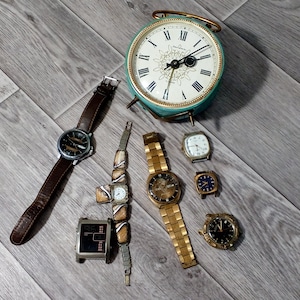 May include: A collection of eight wristwatches and a vintage alarm clock with a teal face and gold accents. The watches are arranged on a wooden floor. The watches have different styles, colours, and materials.