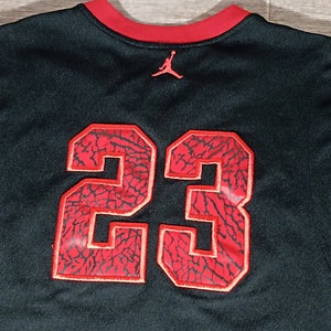 Puede incluir: Camiseta de baloncesto negra con un n&uacute;mero 23 rojo en la espalda. El n&uacute;mero est&aacute; bordeado de blanco y tiene un patr&oacute;n de elefante rojo y negro. La camiseta tiene un cuello rojo y un peque&ntilde;o logotipo Jumpman rojo en la espalda.