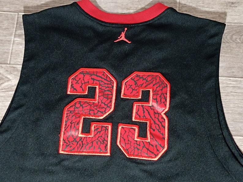 Puede incluir: Camiseta de baloncesto negra con un n&uacute;mero 23 rojo en la espalda. El n&uacute;mero est&aacute; bordeado de blanco y tiene un patr&oacute;n de elefante rojo y negro. La camiseta tiene un cuello rojo y un peque&ntilde;o logotipo Jumpman rojo en la espalda.