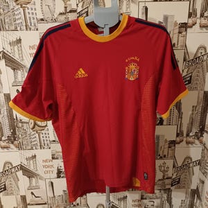 May include: A red Adidas soccer jersey with yellow and navy blue accents. The jersey features the Spanish coat of arms and the word "España" above it. The Adidas logo is visible on the right chest.