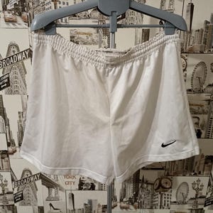 May include: White athletic shorts with an elasticated waistband and a small black Nike swoosh logo. The shorts are hanging on a grey hanger. The shorts are made of a lightweight material.