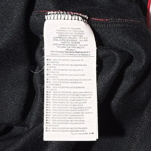 Puede incluir: Una etiqueta blanca con texto negro que detalla el contenido del tejido y el origen de la prenda. La etiqueta dice "Hecho en Tailandia" y enumera la composici&oacute;n del tejido como 100% poli&eacute;ster, excluyendo la decoraci&oacute;n, en varios idiomas.