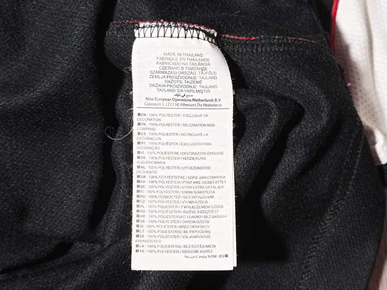 Puede incluir: Una etiqueta blanca con texto negro que detalla el contenido del tejido y el origen de la prenda. La etiqueta dice "Hecho en Tailandia" y enumera la composici&oacute;n del tejido como 100% poli&eacute;ster, excluyendo la decoraci&oacute;n, en varios idiomas.