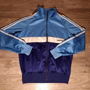 Może przedstawiać: Niebiesko-biało-granatowa kurtka dresowa Adidas z pełnym zamkiem błyskawicznym. Kurtka posiada białe paski na rękawach i logo Adidas na piersi. Posiada wysoki kołnierz oraz prążkowane mankiety i dół.