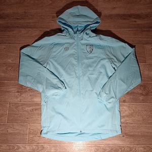 Può includere: Una giacca blu chiaro con cappuccio e chiusura a zip. La giacca ha un logo sul petto sinistro e uno stemma sul petto destro.