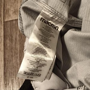 May include: A white tag with black text that reads "macron" and includes information about the product's origin and care instructions. The tag is attached to a gray piece of clothing.