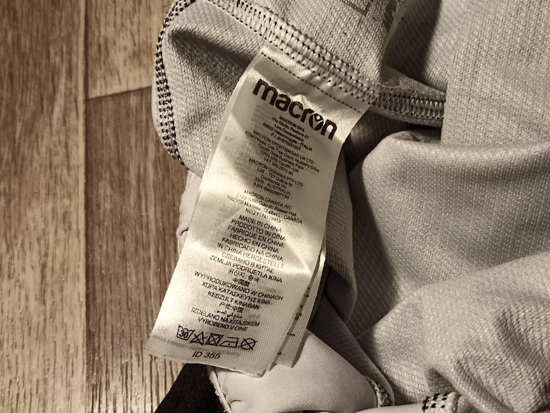 May include: A white tag with black text that reads "macron" and includes information about the product's origin and care instructions. The tag is attached to a gray piece of clothing.