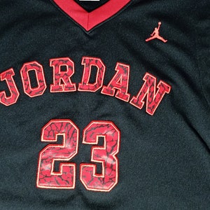 Puede incluir: Camiseta de baloncesto negra con ribetes rojos y la palabra "JORDAN" en rojo con un patr&oacute;n negro y rojo. El n&uacute;mero "23" tambi&eacute;n est&aacute; en rojo con un patr&oacute;n negro y rojo. Un logotipo Jumpman rojo est&aacute; en el lado derecho de la camiseta.