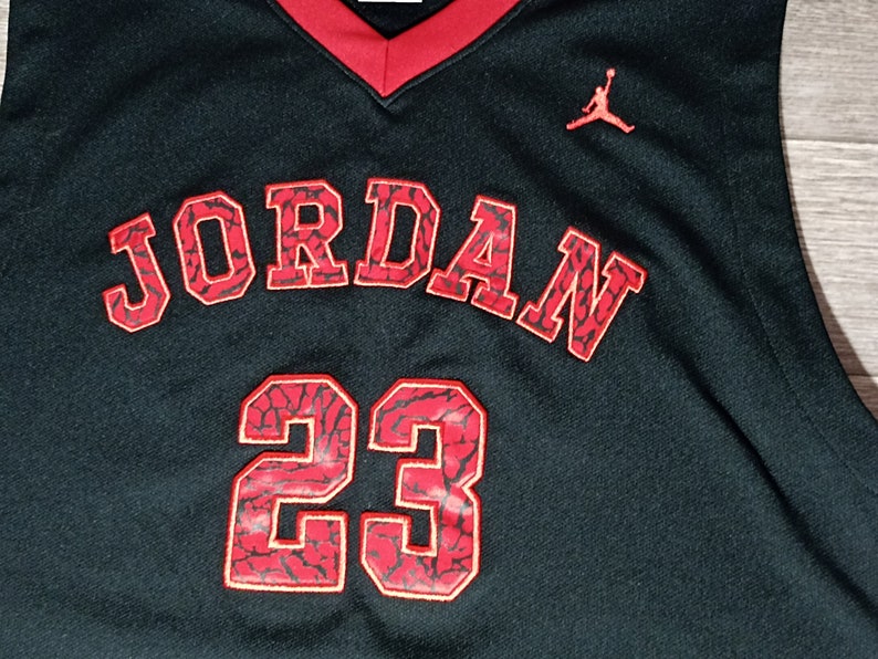 Puede incluir: Camiseta de baloncesto negra con ribetes rojos y la palabra "JORDAN" en rojo con un patr&oacute;n negro y rojo. El n&uacute;mero "23" tambi&eacute;n est&aacute; en rojo con un patr&oacute;n negro y rojo. Un logotipo Jumpman rojo est&aacute; en el lado derecho de la camiseta.