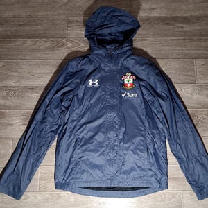 Könnte beinhalten: Eine marineblaue Kapuzenjacke mit Reißverschluss. Die Jacke hat ein weißes Logo auf der linken Brust und ein Teamlogo auf der rechten Brust. Der Text "Sure" ist unter dem Teamlogo gedruckt.