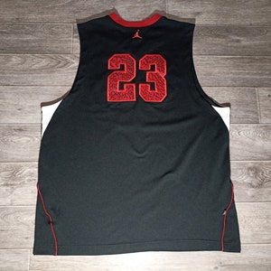 Puede incluir: Camiseta de baloncesto negra con ribetes blancos y detalles rojos. El n&uacute;mero "23" est&aacute; impreso en la espalda en rojo con un patr&oacute;n blanco y negro.