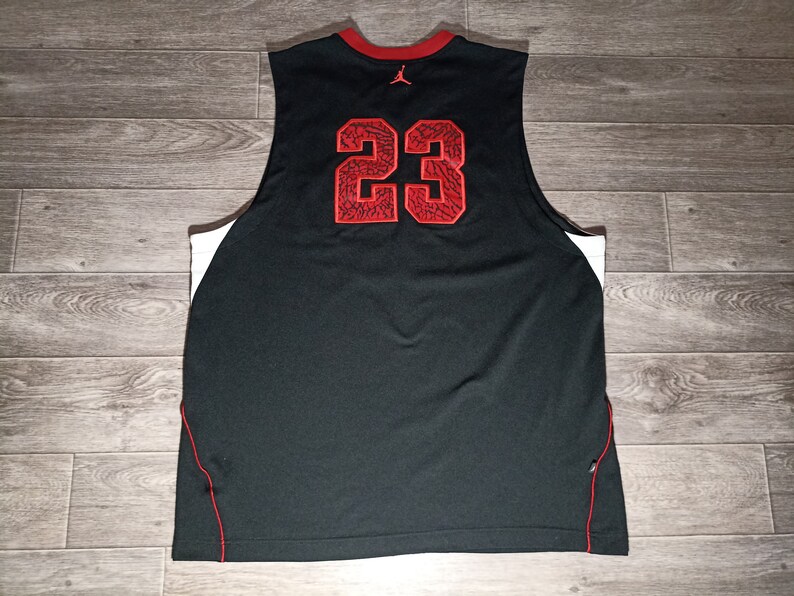 Puede incluir: Camiseta de baloncesto negra con ribetes blancos y detalles rojos. El n&uacute;mero "23" est&aacute; impreso en la espalda en rojo con un patr&oacute;n blanco y negro.