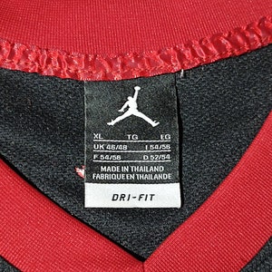 Puede incluir: Camiseta deportiva negra y roja con una etiqueta blanca que dice "XL, UK 46/48, F 54/56, TG, 1 54/56, D 52/54, EG, MADE IN THAILAND, FABRIQUEEN THAILANDE, DRI-FIT".